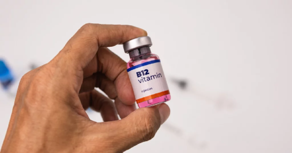 Close-up of a hand holding a vial labeled vitamin B12 injection with a pink liquid inside, against a white background with medical supplies blurred in the back in Sarasota, FL