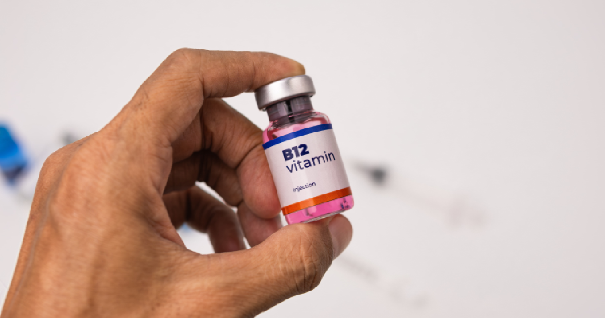 Close-up of a hand holding a vial labeled vitamin B12 injection with a pink liquid inside, against a white background with medical supplies blurred in the back in Sarasota, FL